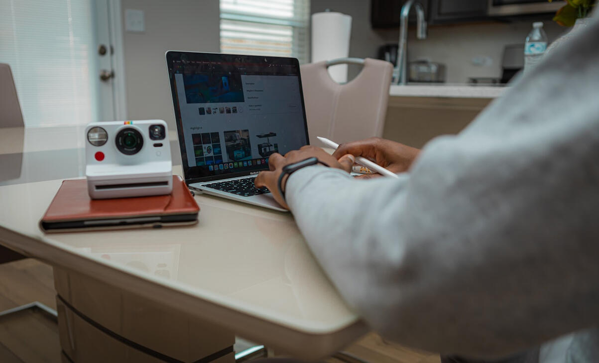 A Black woman sits at a desk working on MacBook while holding an Apple Pen. The image cuts off at her shoulder and focuses on the laptop, her hands, and arms.